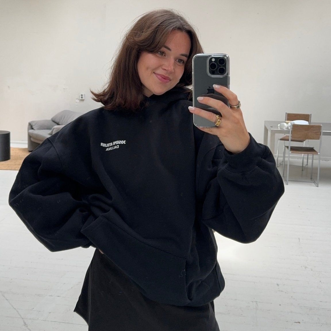 Eine Frau mit schulterlangen braunen Haaren macht ein Selfie im Spiegel in einem hellen, minimalistischen Raum. Sie trägt den DALUMA JOSEPH ATELIER X DALUMA REHAB HOODIE und einen schwarzen Rock und lächelt leicht, während im Hintergrund lässige Möbel zu sehen sind.