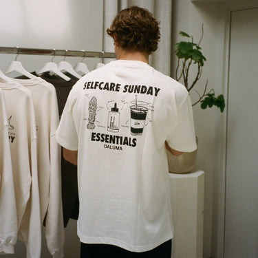 Eine Person mit lockigem Haar trägt das JOSEPH ATELIER X DALUMA SELFCARE SUNDAY TEE von DALUMA, ein übergroßes T-Shirt aus Bio-Baumwolle mit dem Aufdruck SELFCARE SUNDAY ESSENTIALS DALUMA" und Abbildungen von Selbstpflegeprodukten auf dem Rücken. Kleidung und eine Pflanze sind in der Nähe.