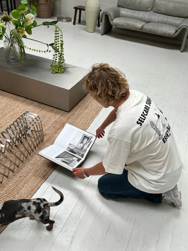Eine Person mit lockigem Haar im DALUMA JOSEPH ATELIER X DALUMA SELFCARE SUNDAY TEE kniet auf einem weißen Boden und schaut auf ein aufgeschlagenes Buch, während ein kleiner Hund auf einem gewebten Teppich neben einem modernen Couchtisch mit einem Pflanzenarrangement spazieren geht.