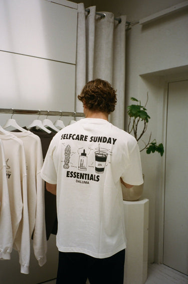 Eine Person mit kurzen braunen Haaren steht in einem Haus und trägt das JOSEPH ATELIER X DALUMA SELFCARE SUNDAY TEE von DALUMA - ein weißes T-Shirt aus Bio-Baumwolle in Übergröße mit Illustrationen zur nachhaltigen Selbstpflege und dem Text SELFCARE SUNDAY ESSENTIALS DALUMA".