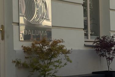 Ein glänzendes Metallschild mit der Aufschrift DALUMA ist an einer hellen Hauswand in der Nähe eines Fensters angebracht. Vor dem Schild stehen zwei getopfte Ahornpflanzen mit grünen und roten Blättern.