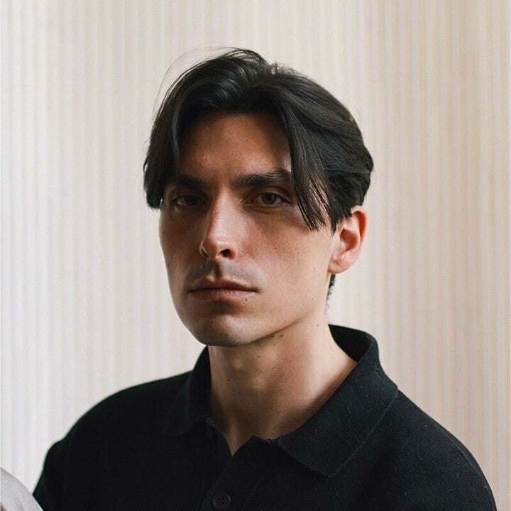 Ein junger Mann mit heller Haut und dunklem, in der Mitte gescheiteltem Haar blickt ernst in die Kamera. Er trägt ein schwarzes Hemd mit Kragen und steht vor einem sanft beleuchteten, beige gestreiften Hintergrund.