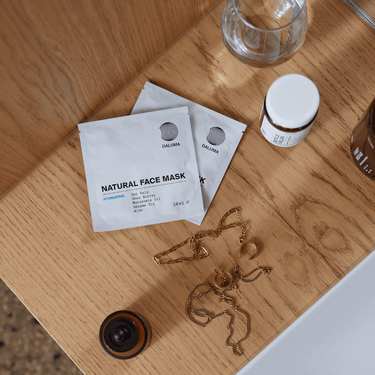 Zwei DALUMA DEEP HYDRATION FEUCHTIGKEITSMASKE Beutel, ein kleines Glas, Goldschmuck und eine braune Flasche mit natürlichen Zutaten sind auf einer Holzfläche neben einem weißen Waschbecken angeordnet.