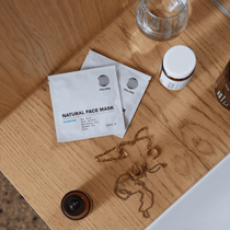 Zwei Päckchen DALUMA DEEP HYDRATION FEUCHTIGKEITSMASKE, ein kleiner Krug, eine goldene Kette und ein Glas ruhen auf Holz neben einem weißen Waschbecken - ideal für den Genuss von Tiefenhydration mit natürlichen Zutaten.