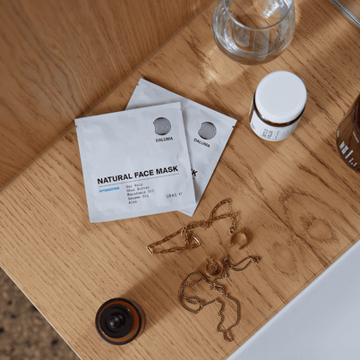 Zwei DALUMA DEEP HYDRATION FEUCHTIGKEITSMASKE Beutel, ein kleines Glas, Goldschmuck und eine braune Flasche mit natürlichen Zutaten sind auf einer Holzfläche neben einem weißen Waschbecken angeordnet.