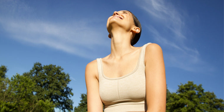 Eine Frau, die ein leichtes Tank-Top trägt, sitzt im Freien, lächelt und legt den Kopf zurück, unter einem klaren blauen Himmel, mit Bäumen im Hintergrund.