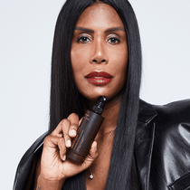 Eine Person mit langen, glatten schwarzen Haaren und rotem Lippenstift hält eine braune Flasche DALUMA FACE RECOVERY MIST GESICHTSSPRAY in die Höhe. Sie trägt eine schwarze Lederjacke vor einem schlichten Hintergrund - perfekt, um Ihren Teint zu schützen.