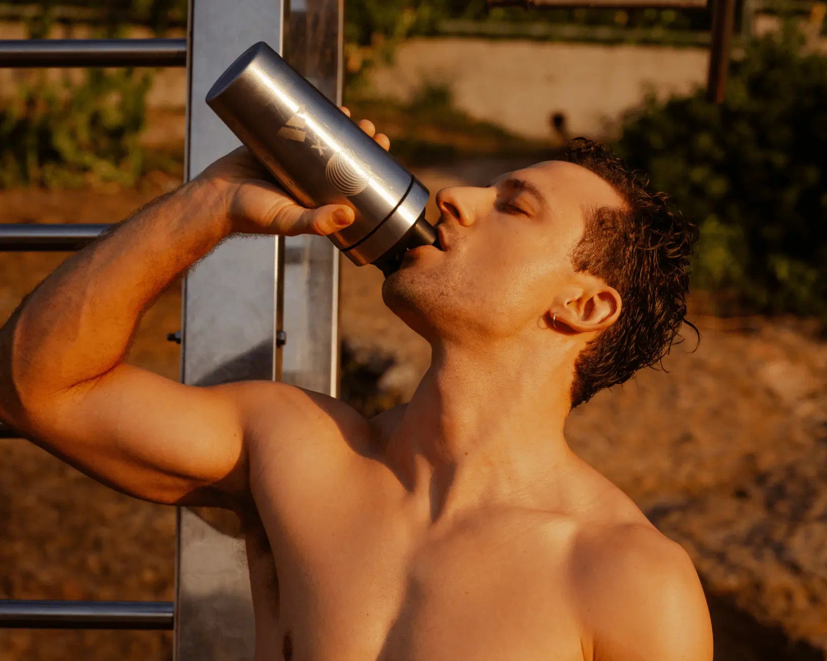 Ein Mann ohne Hemd und mit nassem Haar trinkt aus einer Metallflasche, die im Freien an einer Metallkonstruktion steht, und füllt seinen Eiweißvorrat mit dem VEGAN PERFORMANCE PROTEIN STARTER PACK von DALUMA auf, während das Sonnenlicht einen warmen Schimmer auf seine Haut zaubert.