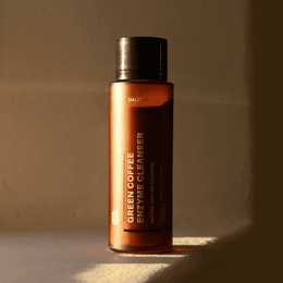 Eine braune Flasche mit der Aufschrift DALUMA GREEN COFFEE CLEANSER ENZYMPEELING steht aufrecht im Sonnenlicht vor einer beigen Wand, wobei warmes Licht und Schatten diese exfolierende Gesichtspflege für natürliche Hautregeneration hervorheben.