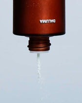 Eine braune Flasche mit der Aufschrift DALUMA, die auf dem Kopf steht, spendet ein feines weißes Pulver von GREEN COFFEE CLEANSER ENZYMPEELING für die exfolierende Gesichtspflege aus ihrer offenen Düse vor einem schlichten Hintergrund.