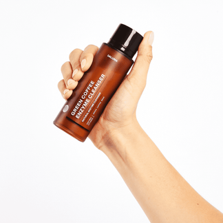Eine Hand hält eine braune Flasche mit der Aufschrift GREEN COFFEE CLEANSER ENZYMPEELING von DALUMA vor einem weißen Hintergrund - ideal für die exfolierende Hautpflege und das sanfte Peeling zur Unterstützung der natürlichen Regeneration.