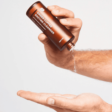 Eine Hand gießt DALUMA GREEN COFFEE CLEANSER ENZYMPEELING aus einer braunen Flasche auf eine geöffnete Handfläche und hebt das sanfte Peeling zur natürlichen Regeneration vor einem schlichten weißen Hintergrund hervor.