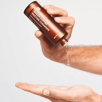 Eine Person schüttet Pulver aus einer braunen DALUMA-Flasche mit der Aufschrift GREEN COFFEE CLEANSER ENZYMPEELING auf ihre Handfläche und hebt das Peeling-Gesichtspflegeprodukt vor einem schlichten weißen Hintergrund hervor.