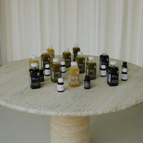 Auf einem runden Steintisch stehen verschiedene kleine Flaschen und Behälter mit Flüssigkeiten in grüner, gelber und dunkler Farbe, die vor einem hellen Vorhang stehen.