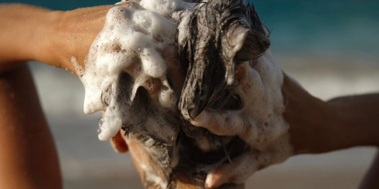 Eine Person wäscht sich die Haare mit Shampoo und schäumt dabei mit beiden Händen dicke, schaumige Blasen auf. Der Hintergrund ist unscharf und suggeriert eine Umgebung im Freien, möglicherweise in der Nähe des Meeres oder Ozeans.