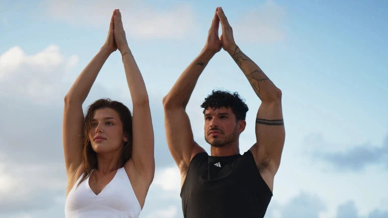 Eine Frau und ein Mann üben am Strand Yoga, indem sie mit über den Kopf gehobenen Armen in Baumhaltung stehen. Die Frau trägt weiße Sportkleidung, der Mann trägt schwarz. Im Hintergrund sind das Meer und ein bewölkter Himmel zu sehen.