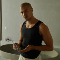 Ein Mann in einem schwarzen Tank-Top steht in einem minimalistischen Badezimmer und hält die HYDRATING ESSENTIALS von DALUMA in der Hand, wobei er einen neutralen Ausdruck trägt. Im Hintergrund sind eine runde Badewanne und Toilettenartikel zu sehen.