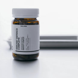 Eine braune Glasflasche mit weißem Deckel und einem Etikett mit der Aufschrift IMMUN DEFENCE COMPLETE KOMPLEX von DALUMA steht auf einer weißen Fläche, perfekt zum Immunsystem stärken mit antimikrobieller Wirkung; im Hintergrund ist ein unscharfes Tablett zu sehen.