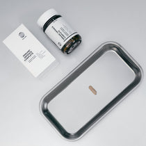 Eine einzelne Kapsel auf einem Metalltablett neben einer weißen DALUMA IMMUN DEFENCE COMPLETE KOMPLEX-Schachtel und einem Pillenfläschchen, die den Knoblauchextrakt zur Stärkung des Immunsystems hervorheben, vor einem hellen Hintergrund.