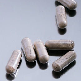 Sieben grau marmorierte IMMUN DEFENCE COMPLETE KOMPLEX Kapseln von DALUMA liegen verstreut auf einer reflektierenden hellgrauen Oberfläche - perfekt für die Stärkung Ihres Immunsystems.