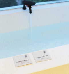 Eine Badewanne wird über einen schwarzen Wasserhahn gefüllt. Auf dem weißen Sims liegen zwei DALUMA 2x MASK BUNDLE Pakete nebeneinander. Im Hintergrund erscheint ein Fenster.
