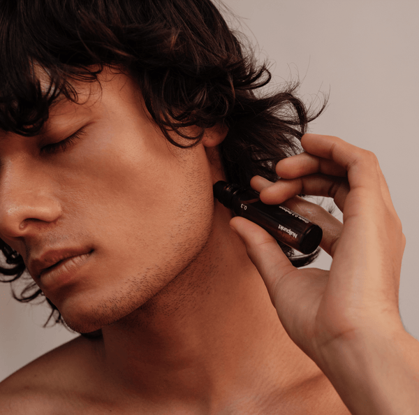 Eine Person mit mittellangem, dunklem Haar trägt vor einem neutralen Hintergrund das Serum COMPLETE COLLECTION von Nullpunkt aus einer Tropfflasche mit geschlossenen Augen sanft im Nacken auf.