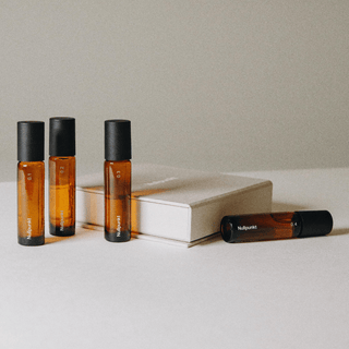 Die Nullpunkt COMPLETE COLLECTION von Nullpunkt zeigt vier Braunglas-Rollerflaschen mit schwarzen Verschlüssen, die neben einem geschlossenen beigen Buch auf weißem Untergrund und neutralem Hintergrund stehen; drei Flaschen stehen aufrecht, eine liegt auf der Seite.