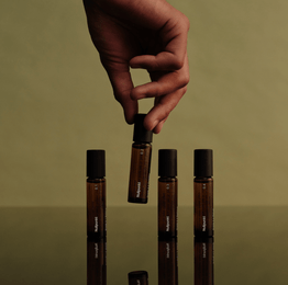 Eine Hand hält eine von vier NULLPUNKT COMPLETE COLLECTION Braunglas-Rollerflaschen von Nullpunkt mit schwarzer Kappe, die auf einer spiegelnden Fläche vor olivgrünem Hintergrund angeordnet sind.
