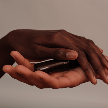 Zwei Hände halten sanft eine kleine, dunkle Flasche aus der NULLPUNKT COMPLETE COLLECTION von Nullpunkt vor einem neutralen Hintergrund, der an Achtsamkeit und Ruhe erinnert.