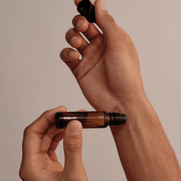 Vor einem neutralen Hintergrund hält eine Person einen kleinen Braunglas-Roll-on-Flakon aus der Nullpunkt Complete Collection von Nullpunkt und entfernt mit der anderen Hand vorsichtig die schwarze Kappe.