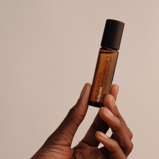 Eine Hand hält eine kleine Braunglas-Tropfflasche mit einer schwarzen Kappe vor einem beigen Hintergrund. Auf dem Etikett steht "NULLPUNKT GRADE 0.1 RELIEVE" von Nullpunkt, was diese minimalistische Hautpflegeszene noch interessanter macht.