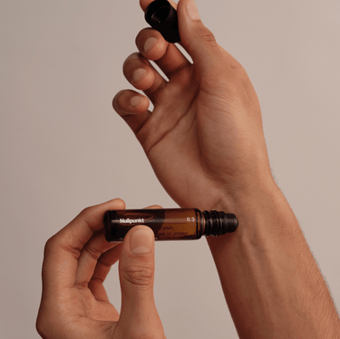 Eine Person hält eine NULLPUNKT GRADE 0.1 RELIEVE Braunglas-Rollerflasche von Nullpunkt und schraubt die schwarze Kappe vor einem einfarbig beigen Hintergrund ab.