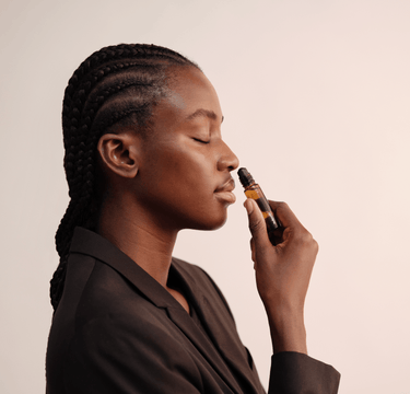 Eine Frau mit geflochtenem Haar und geschlossenen Augen hält einen Nullpunkt GRADE 0.1 RELIEVE Aromatherapie-Ölroller an ihre Nase, als ob sie daran riechen würde, während sie einen schwarzen Blazer vor einem hellen Hintergrund trägt.