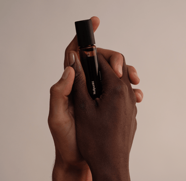 Zwei Hände - eine hellere und eine dunklere - halten sanft den NULLPUNKT GRADE 0.2 RELEASE, einen Parfümflakon aus Braunglas mit schwarzem Verschluss von Nullpunkt, vor einem neutralen beigen Hintergrund.