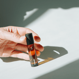 Eine Hand hält einen Flakon NULLPUNKT GRADE 0.3 REFOCUS von Nullpunkt aus Braunglas mit schwarzem Verschluss, der im hellen Sonnenlicht einen scharfen Schatten auf eine weiße Oberfläche wirft.
