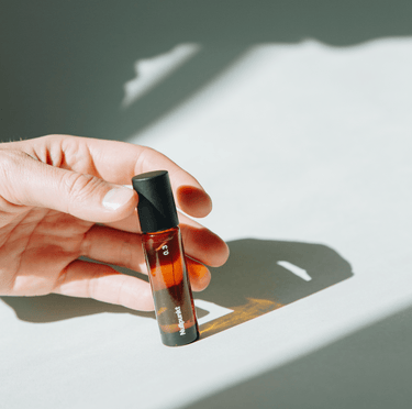 Eine Hand hält einen Flakon NULLPUNKT GRADE 0.3 REFOCUS von Nullpunkt aus Braunglas mit schwarzem Verschluss, der im hellen Sonnenlicht einen scharfen Schatten auf eine weiße Oberfläche wirft.