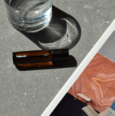 Ein kleines Braunglasfläschchen mit der Aufschrift NULLPUNKT GRADE 0.3 REFOCUS von Nullpunkt steht auf einer grauen Fläche neben einem halbvollen Glas Wasser und einer bunten Magazinseite mit abstrakten Mustern und einem beigen Stuhl.