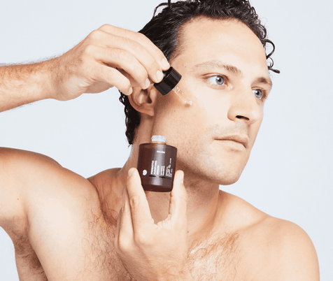 Ein hemdsärmeliger Mann mit kurzem, lockigem Haar trägt DALUMA PREBIOTIC PURIFYING GESICHTSSERUM aus einer kleinen braunen Flasche mit einer Pipette auf seine Wange auf, vor einem schlichten hellen Hintergrund.