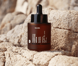 Ein Tropffläschchen aus Braunglas mit der Aufschrift DALUMA PREBIOTIC PURIFYING GESICHTSSERUM ruht unter natürlichem Licht auf strukturierten beigen Steinen und unterstützt die Hautbarriere mit probiotischen Kulturen zur effektiven Reduzierung von Unreinheiten.