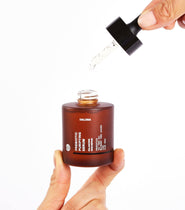 Eine Hand hält eine braune Glasflasche mit der Aufschrift DALUMA PREBIOTIC PURIFYING GESICHTSSERUM, reich an probiotischen Bakterienkulturen, während eine andere Hand das Serum aus einer Pipette über der Flasche vor einem weißen Hintergrund ausgibt.