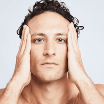 Ein Mann mit nassem Haar und feuchter Haut hält sich das Gesicht und blickt in die Kamera vor einem hellen Hintergrund, der die Wirkung von DALUMA PREBIOTIC PURIFYING GESICHTSSERUM hervorhebt.