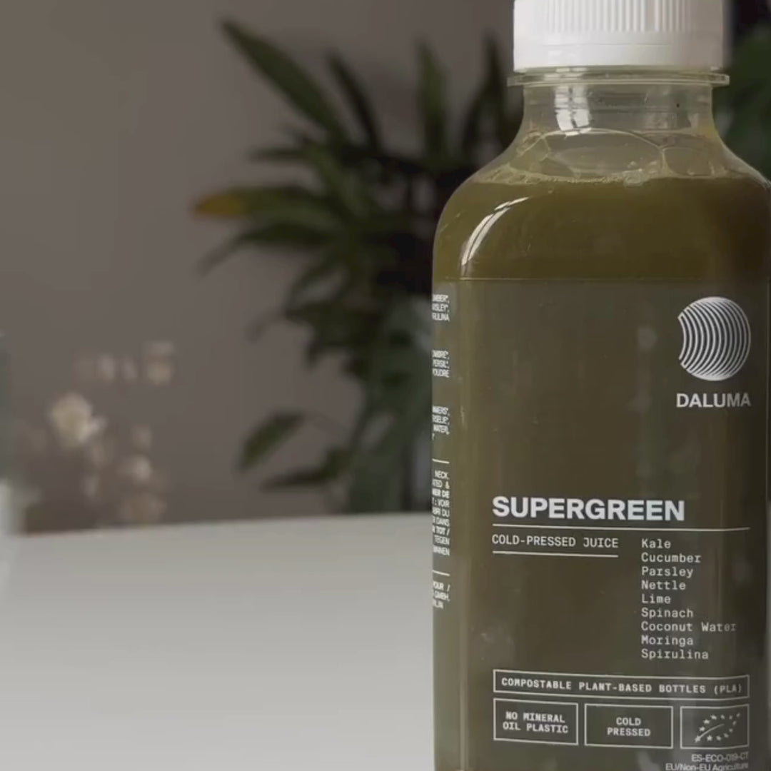 Eine Packung Daluma SUPERGREEN GREEN JUICE 6 PACK steht auf einem Tisch, auf dem die Zutaten auf dem Etikett angegeben sind. Eine Blattpflanze ist unscharf im Hintergrund zu sehen.