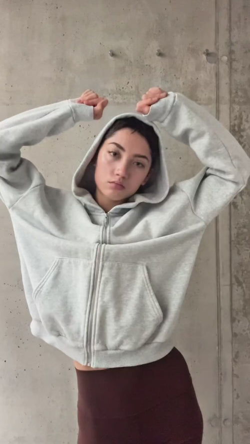 Eine Person, die einen hellgrauen Kapuzenpulli mit aufgesetzter Kapuze trägt, steht an einer Betonwand, hebt die Arme und blickt mit neutralem Blick direkt in die Kamera.