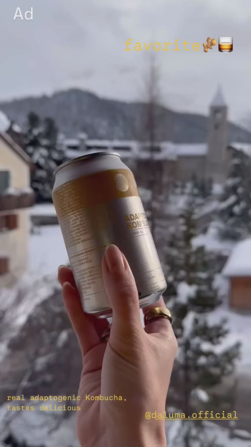 Eine Hand mit lackierten Nägeln hält eine Dose mit adaptogenem Kombucha vor einer verschneiten Dorfszene mit Bergen und einer Kirche im Hintergrund. Der Text auf dem Bild hebt den Kombucha als Lieblingsgetränk hervor.