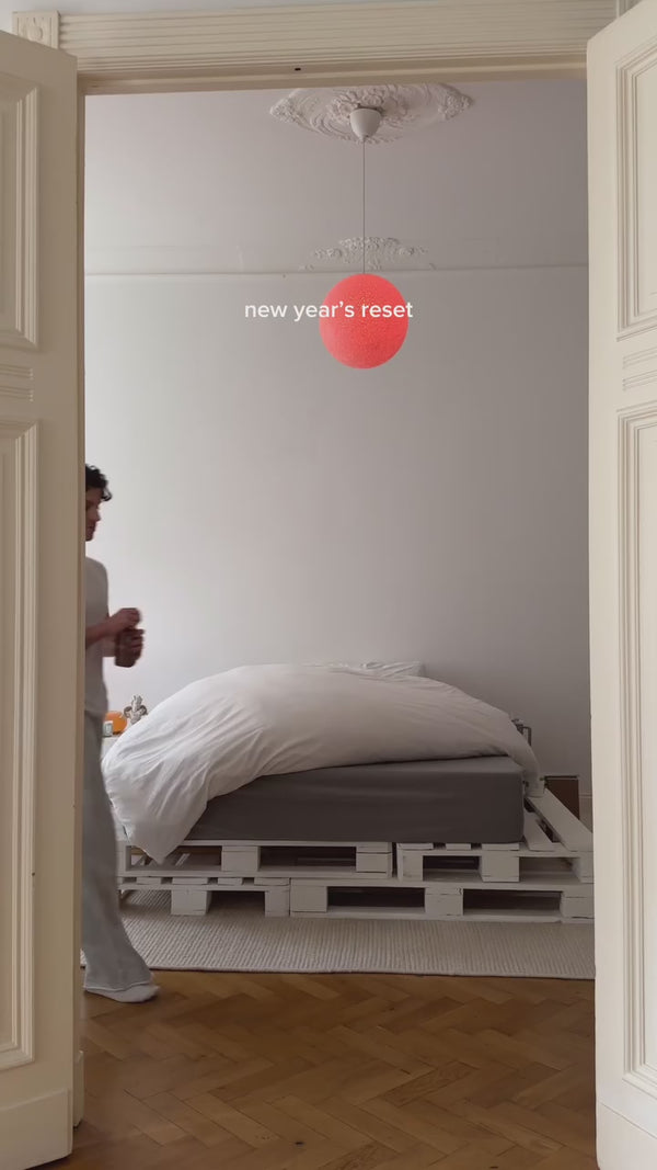 Eine Person betritt ein minimalistisches Schlafzimmer mit einem Bett auf einem weißen Palettenrahmen, weißer Bettwäsche und einer einfachen Leuchte. Text auf dem Bild liest neue Jahre zurückgesetzt.