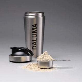 Eine Shakerflasche aus rostfreiem Stahl mit der Aufschrift DALUMA, deren schwarzer Deckel abgenommen ist, befindet sich hinter einer Metallschaufel, die mit Proteinpulver gefüllt ist, das auf eine graue Fläche verschüttet wird.