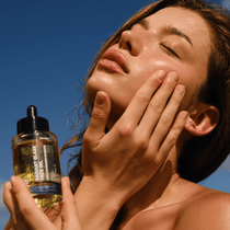 Eine Frau trägt FACE GLOW DUO von DALUMA mit einer Tropfflasche auf ihre Wange auf und sonnt sich. Angereichert mit Granatapfel Extrakt, sieht ihre Haut unter dem klaren blauen Himmel strahlend und gesund aus.