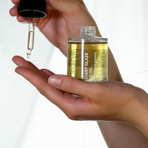Eine Person hält eine Glasflasche mit DALUMA RADIANT GLAZE GESICHTSÖL in der einen Hand und gibt mit einer Pipette Tropfen auf ihre andere Handfläche. Der Hintergrund ist hell und schlicht.
