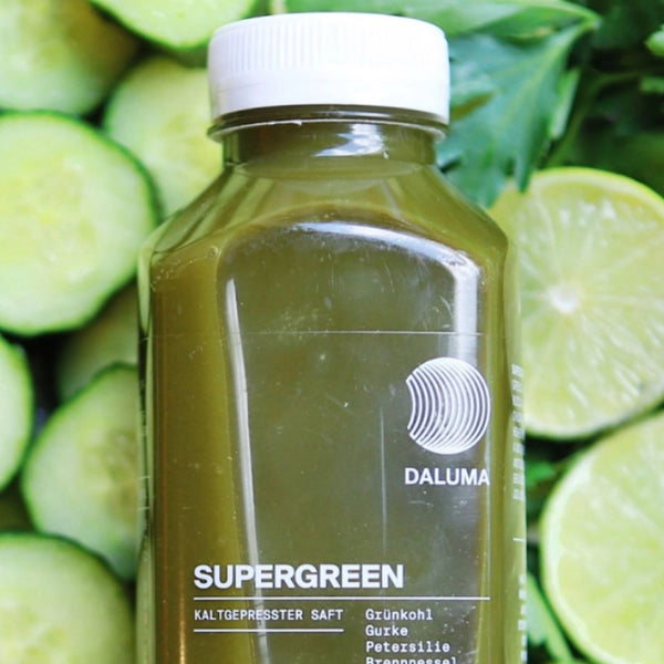 Eine Flasche des kaltgepressten Saftes Supergreen von Daluma wird in Nahaufnahme gezeigt, mit geschnittenen Gurken und Blattgemüse im Hintergrund. Der Saft hat eine grüne Farbe und eine weiße Kappe.