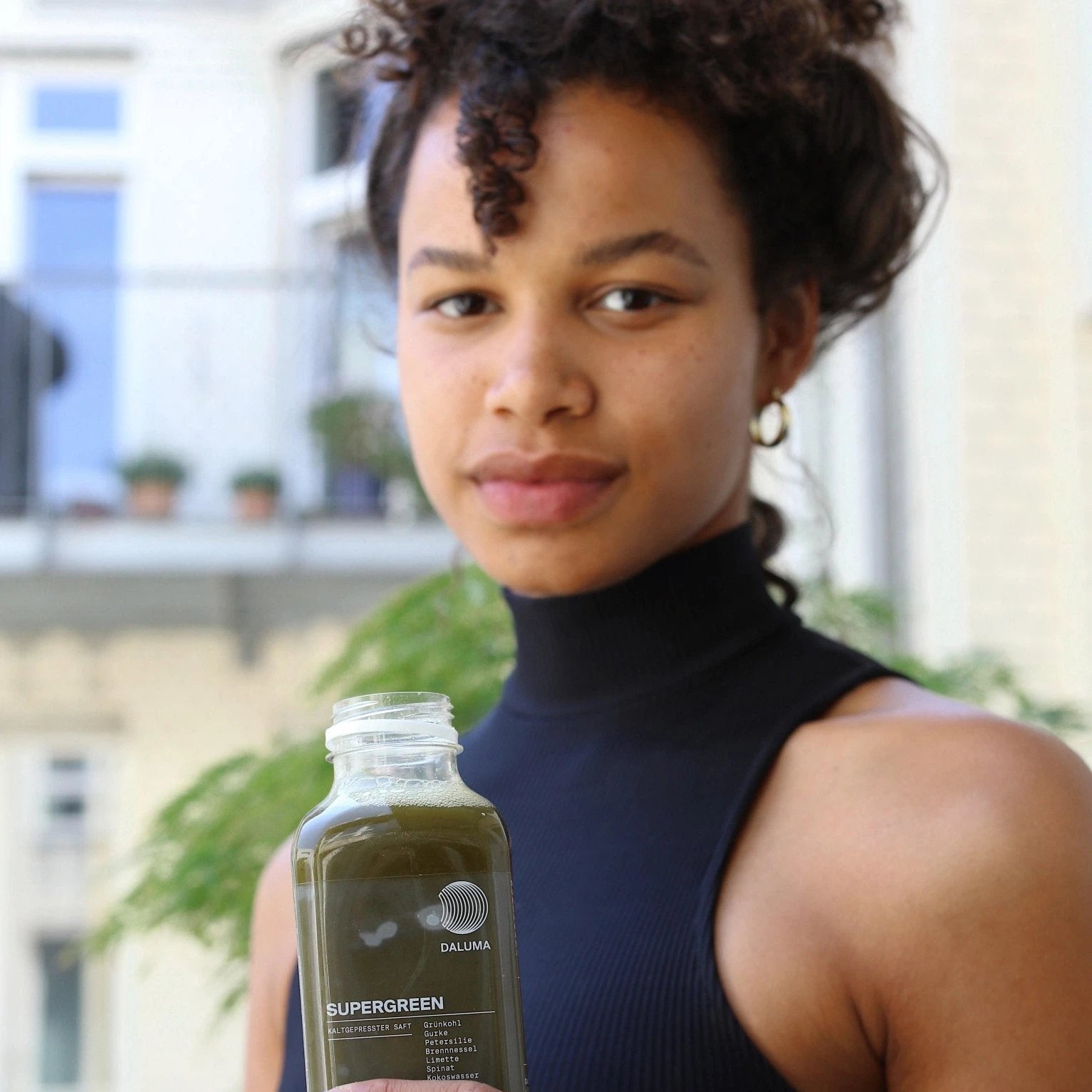 Eine Person mit lockigem Haar in einem ärmellosen schwarzen Oberteil hält eine durchsichtige Flasche DALUMA SUPERGREEN GREEN JUICE 6 PACK und steht im Freien mit Pflanzen und einem Gebäude, das diesen Bio-Kaltgepresster Gemüsesaft hervorhebt.
